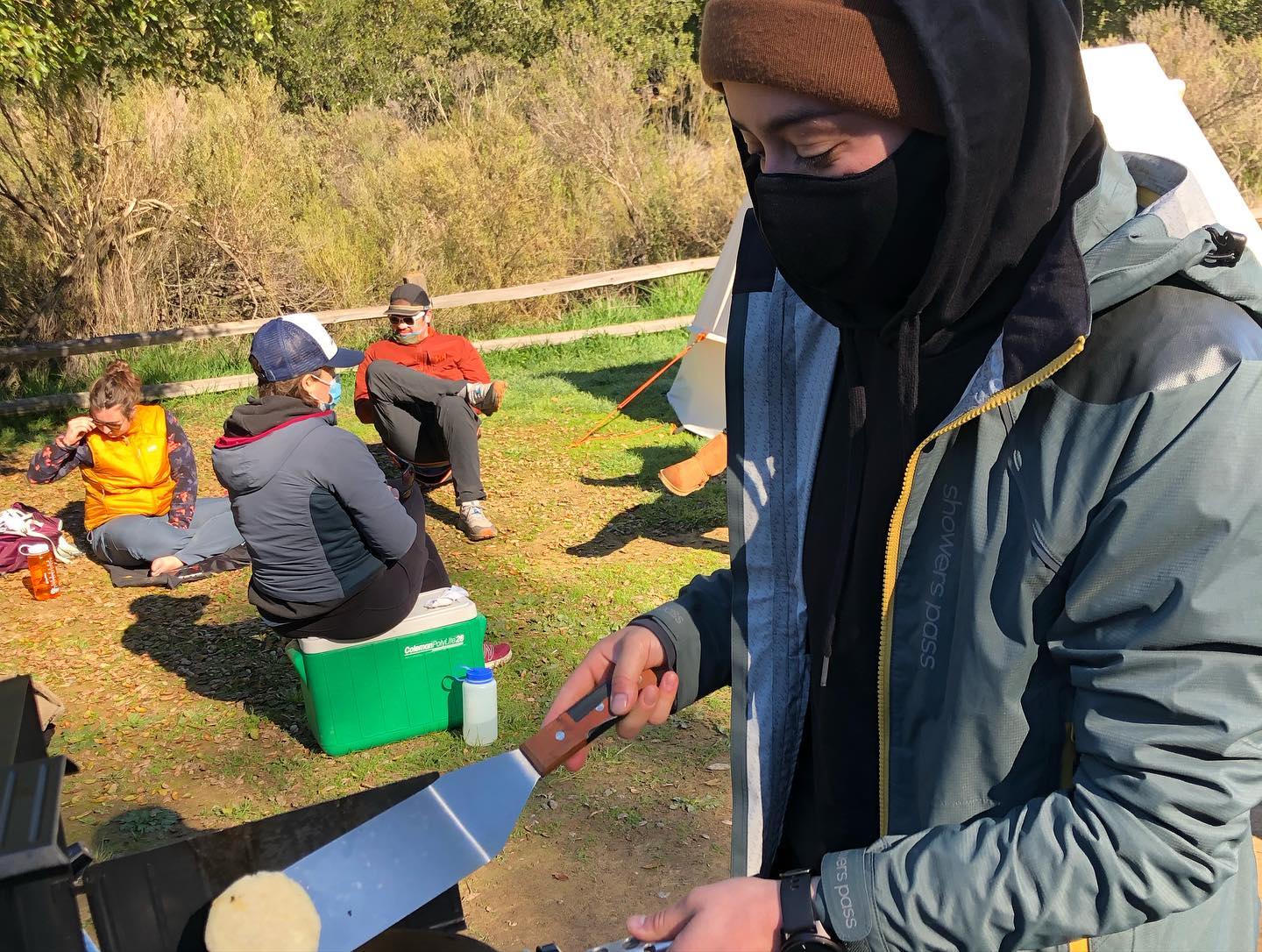 A person bundled up against the cold flips pancakes on a camp stove