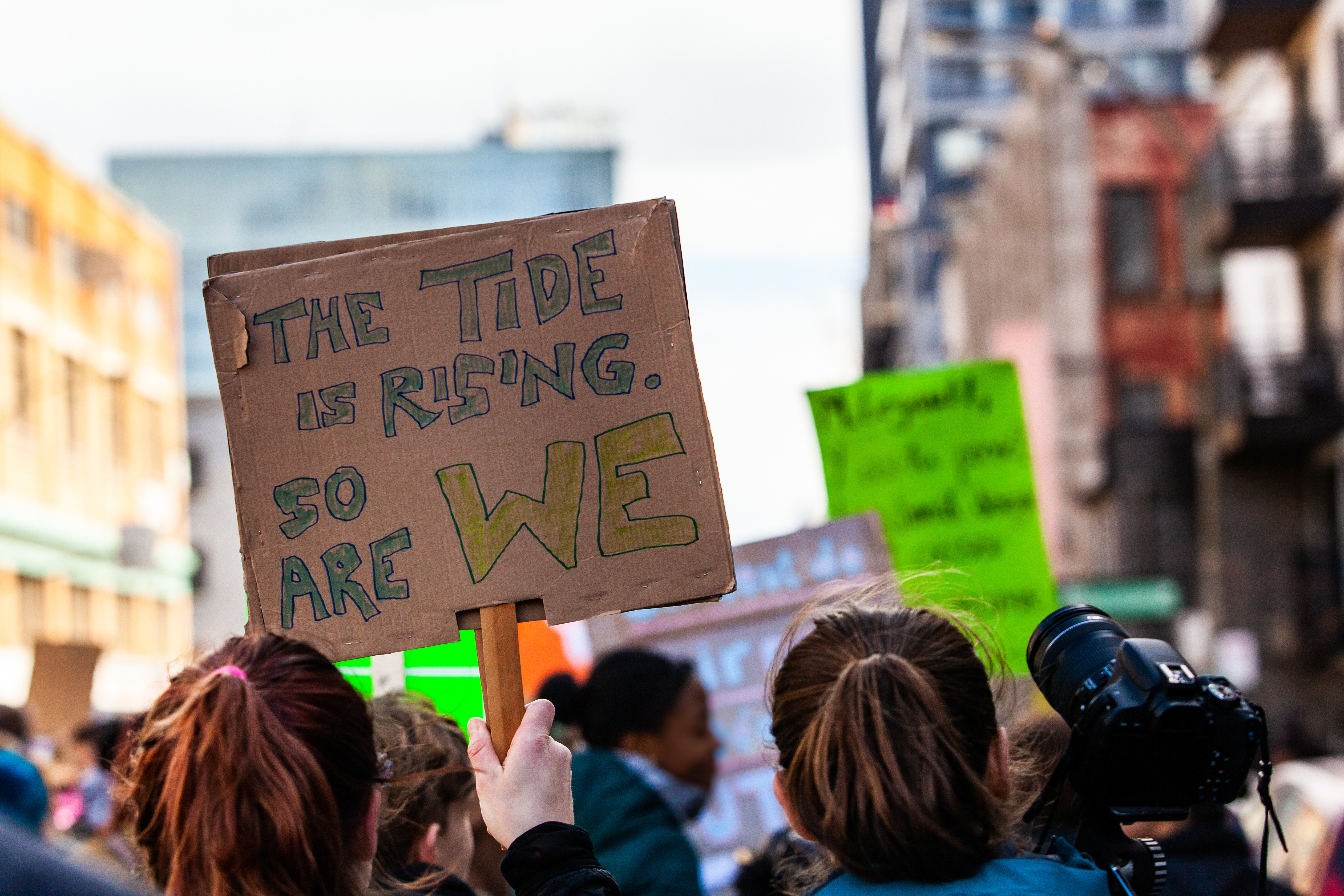 A sign being held at a protest reads "the tide is rising, so are we."