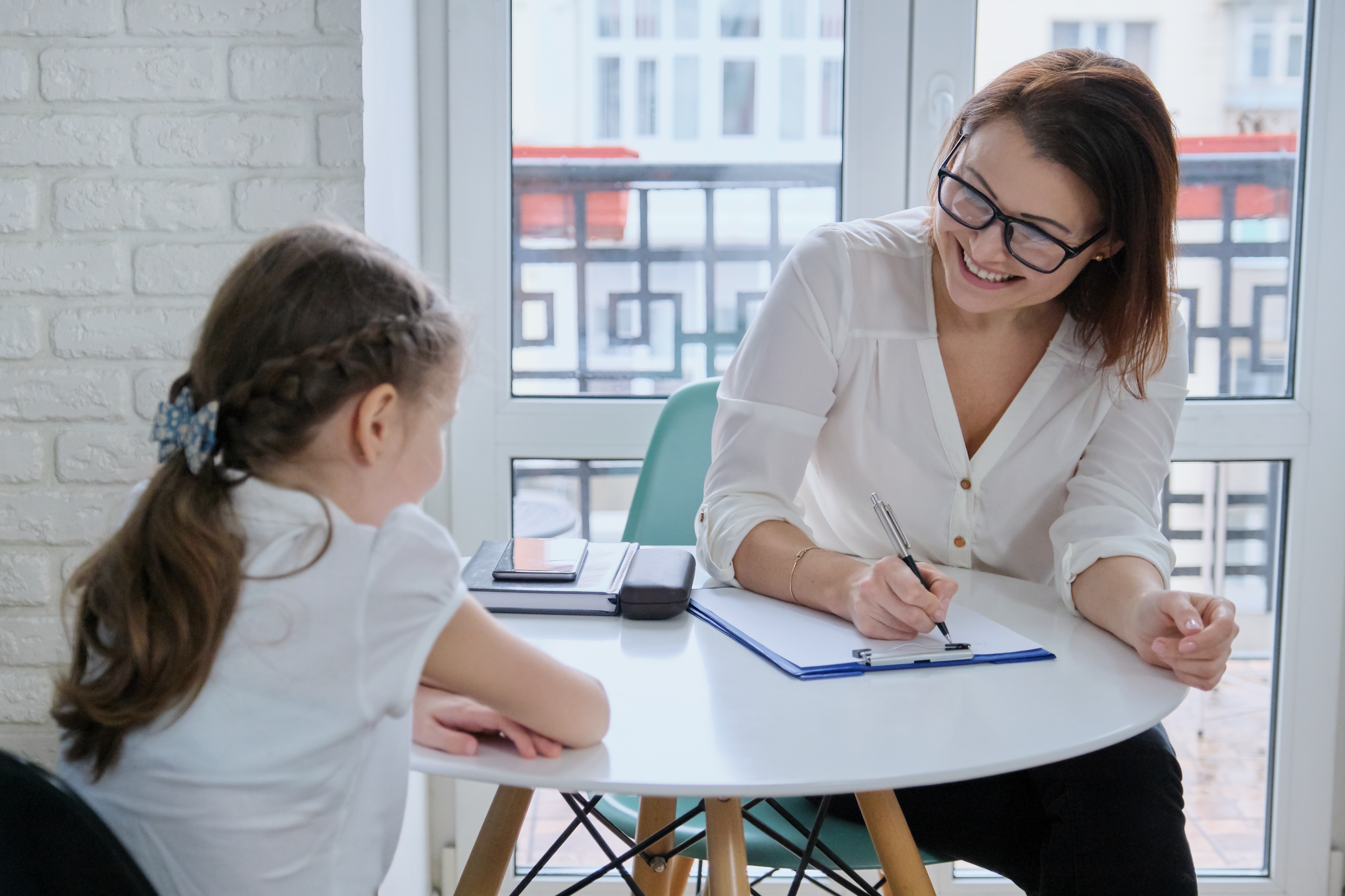 An adult sits with a child at a table holding a clipboard