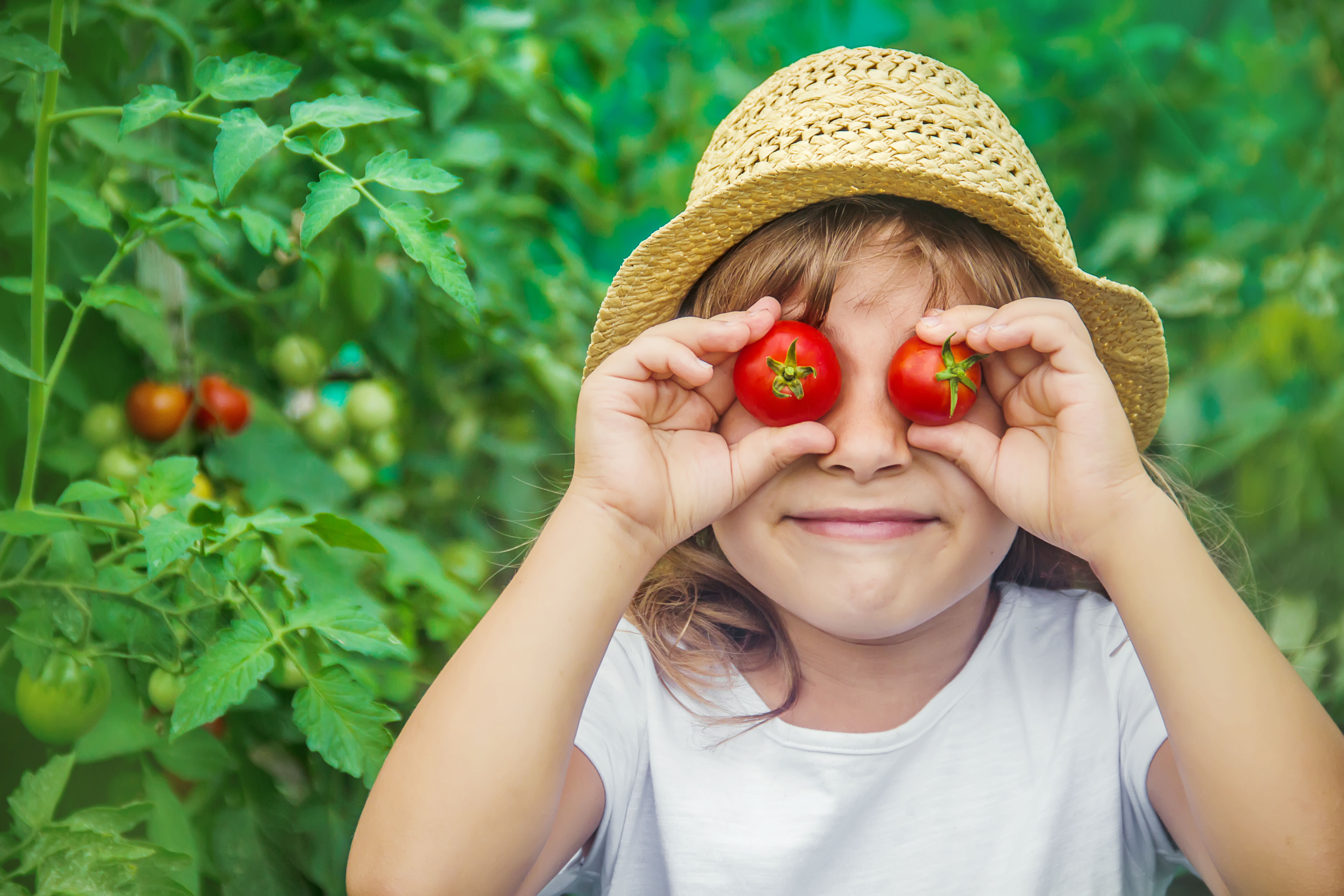 A child holds up tomatoes to her eyes and smiles into the camera. 
