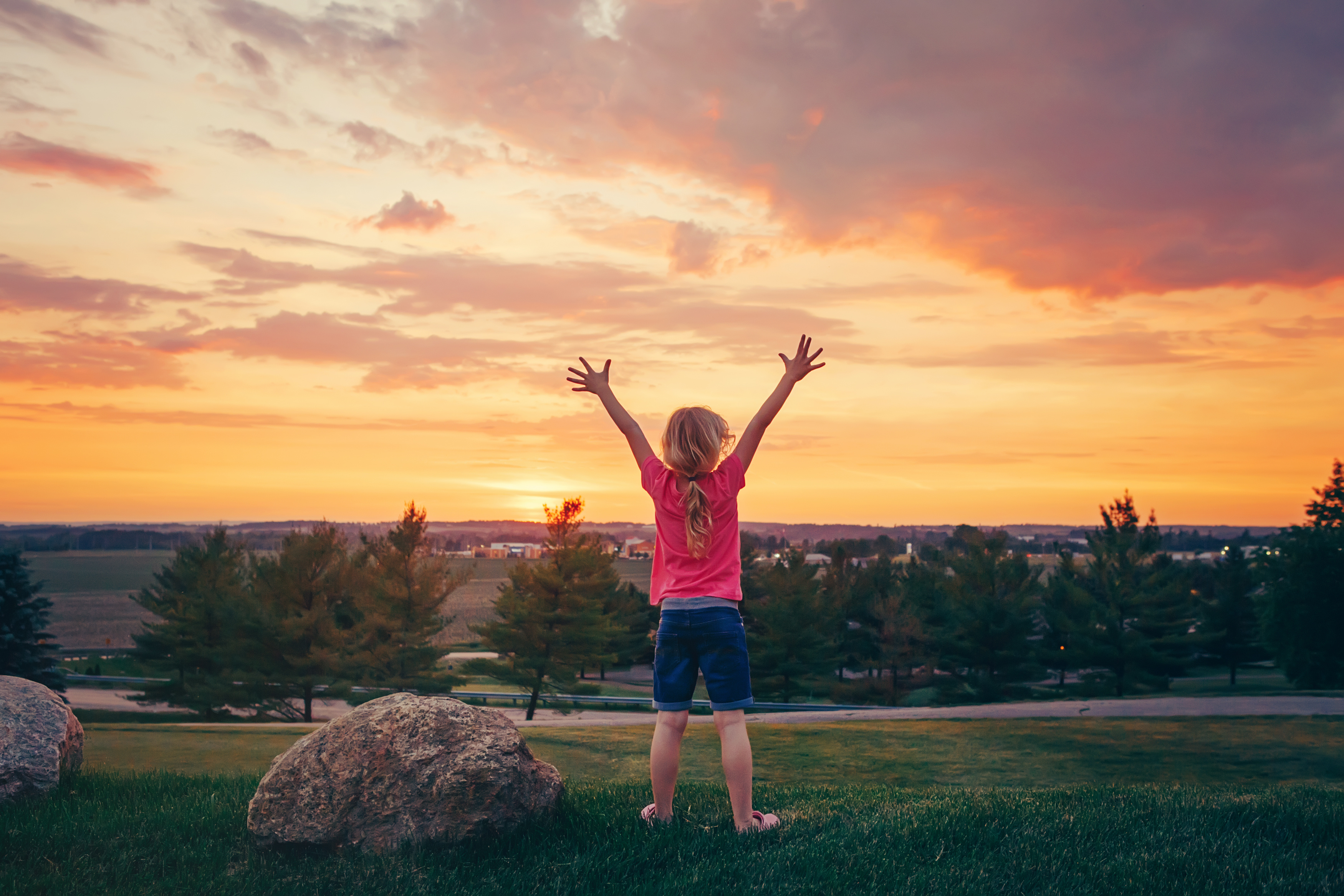 A young child with a long ponytail stands in front of a sunset with her arms raised over her head.