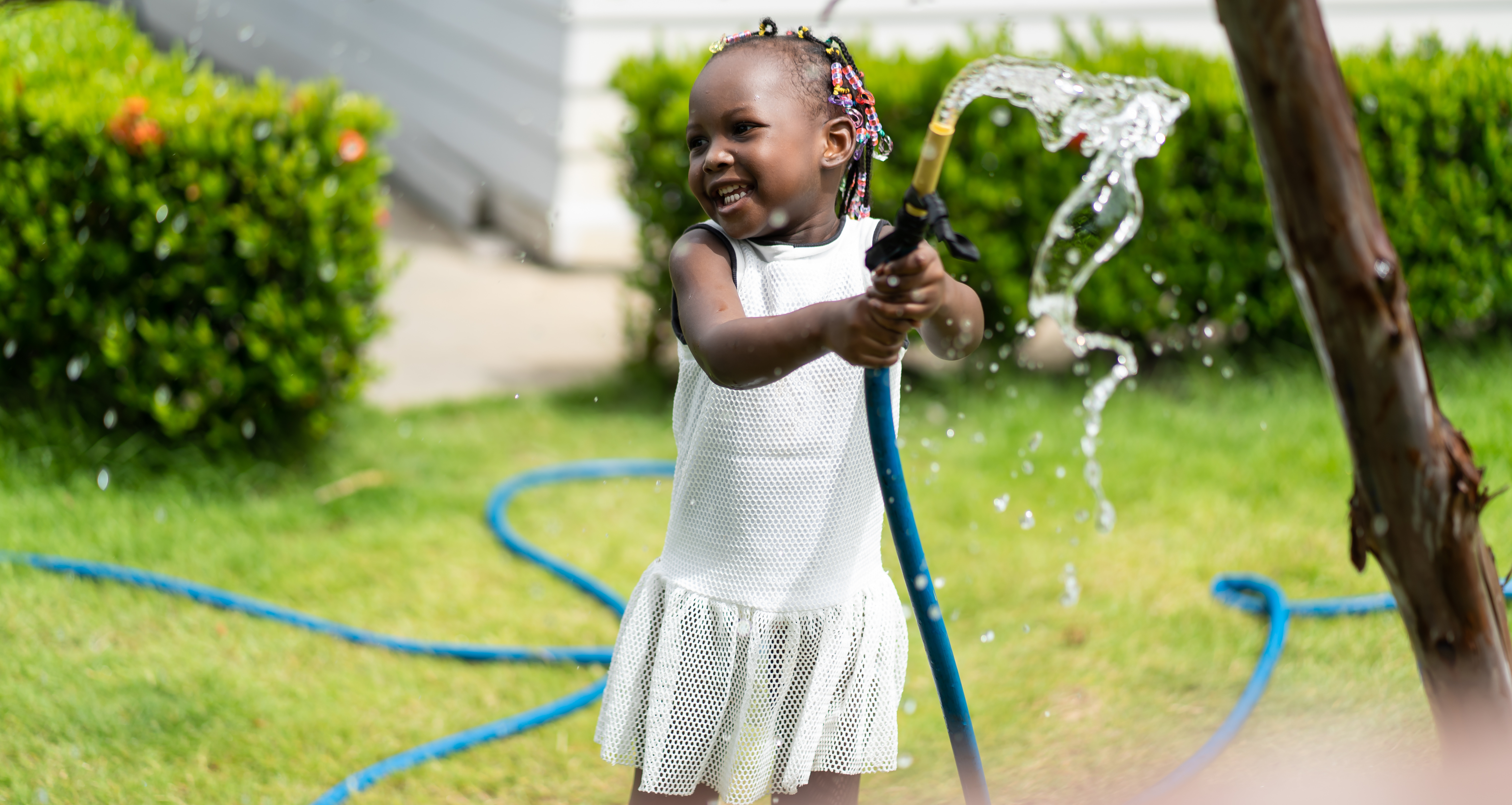 A child holds a hose in the middle of a lawn and smiles