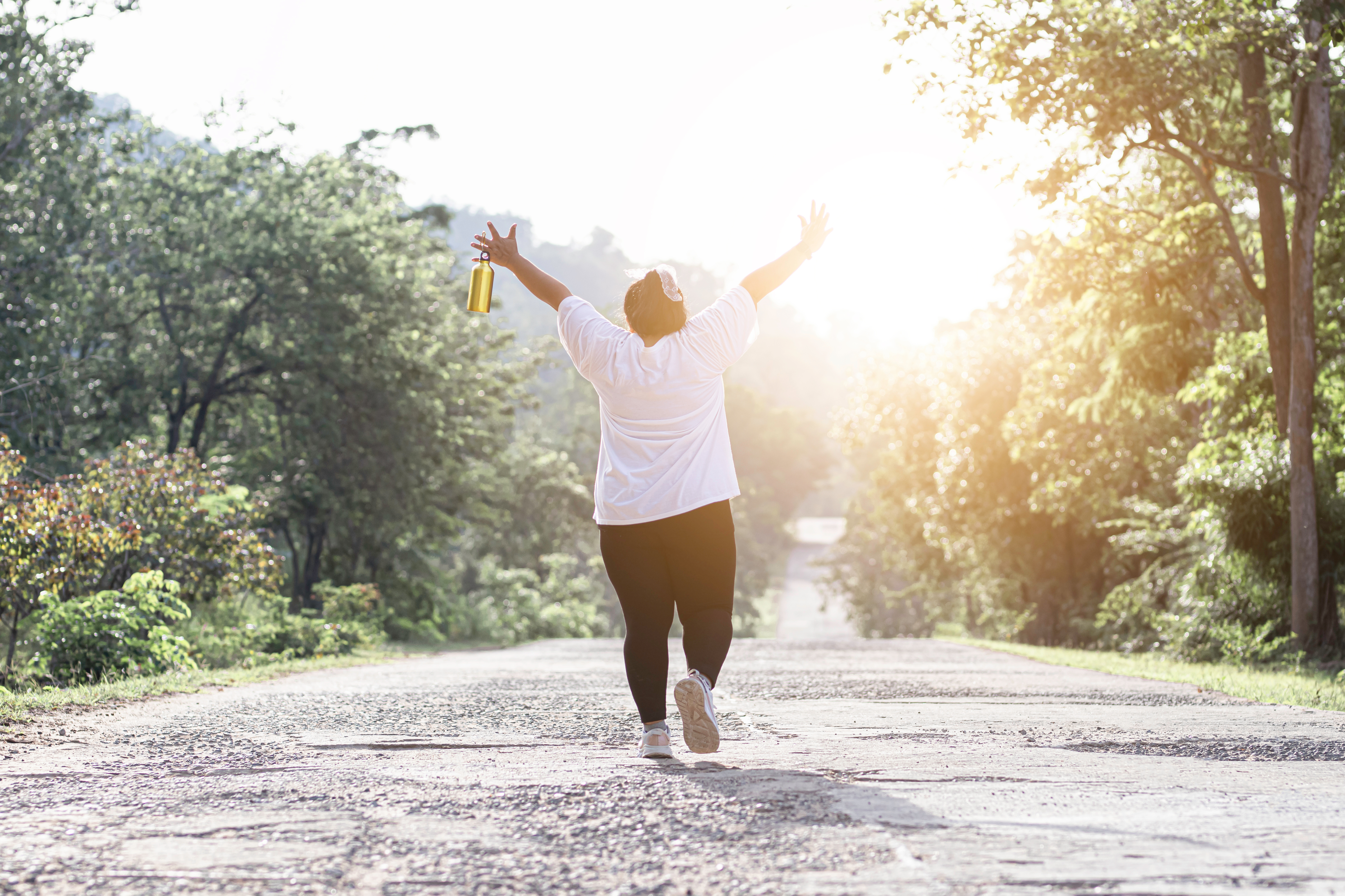 A young woman with her hair tied up walks down a sunny path with her back to the camera