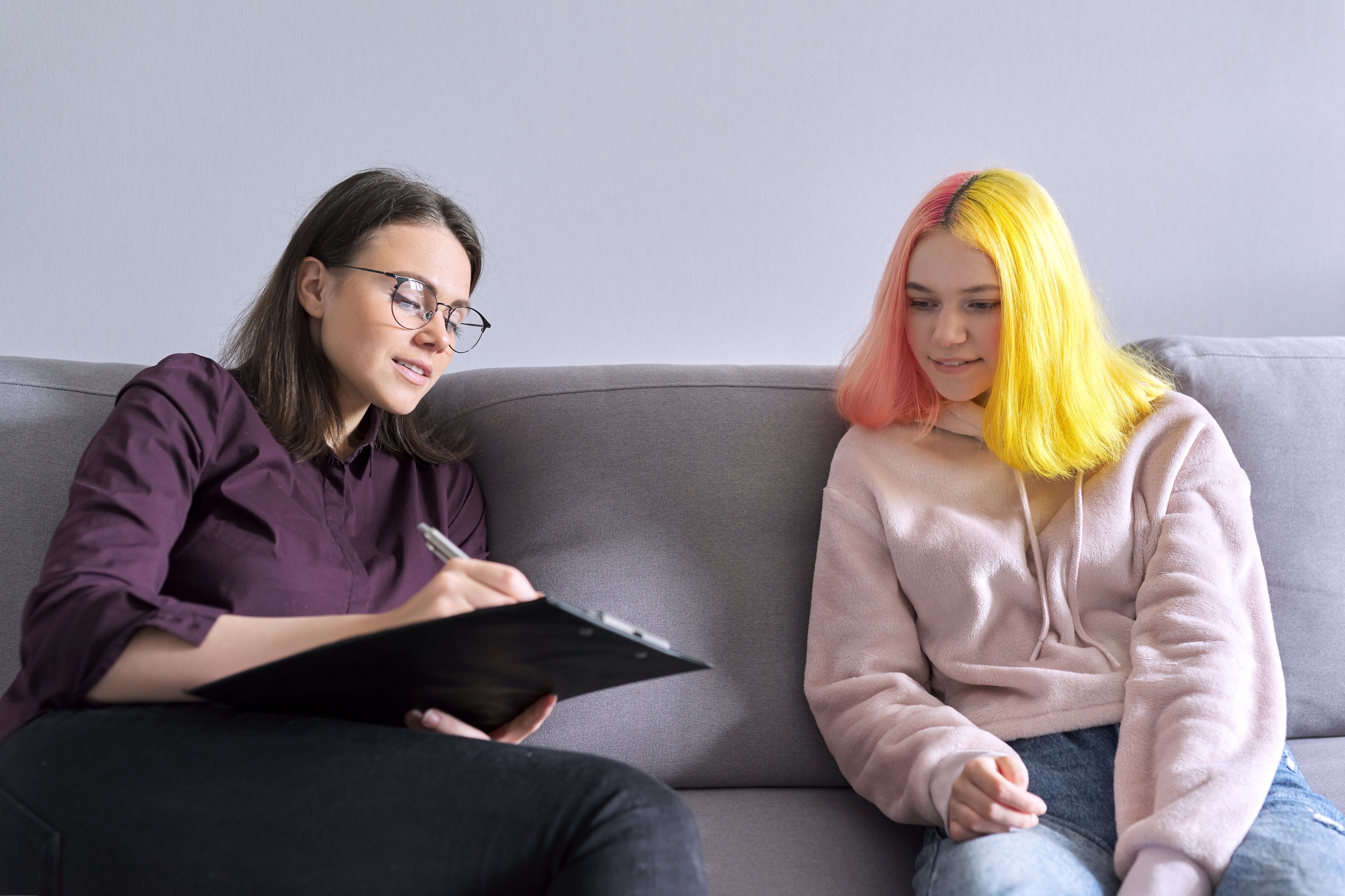 A young person sits next to an adult on a couch who is taking notes 