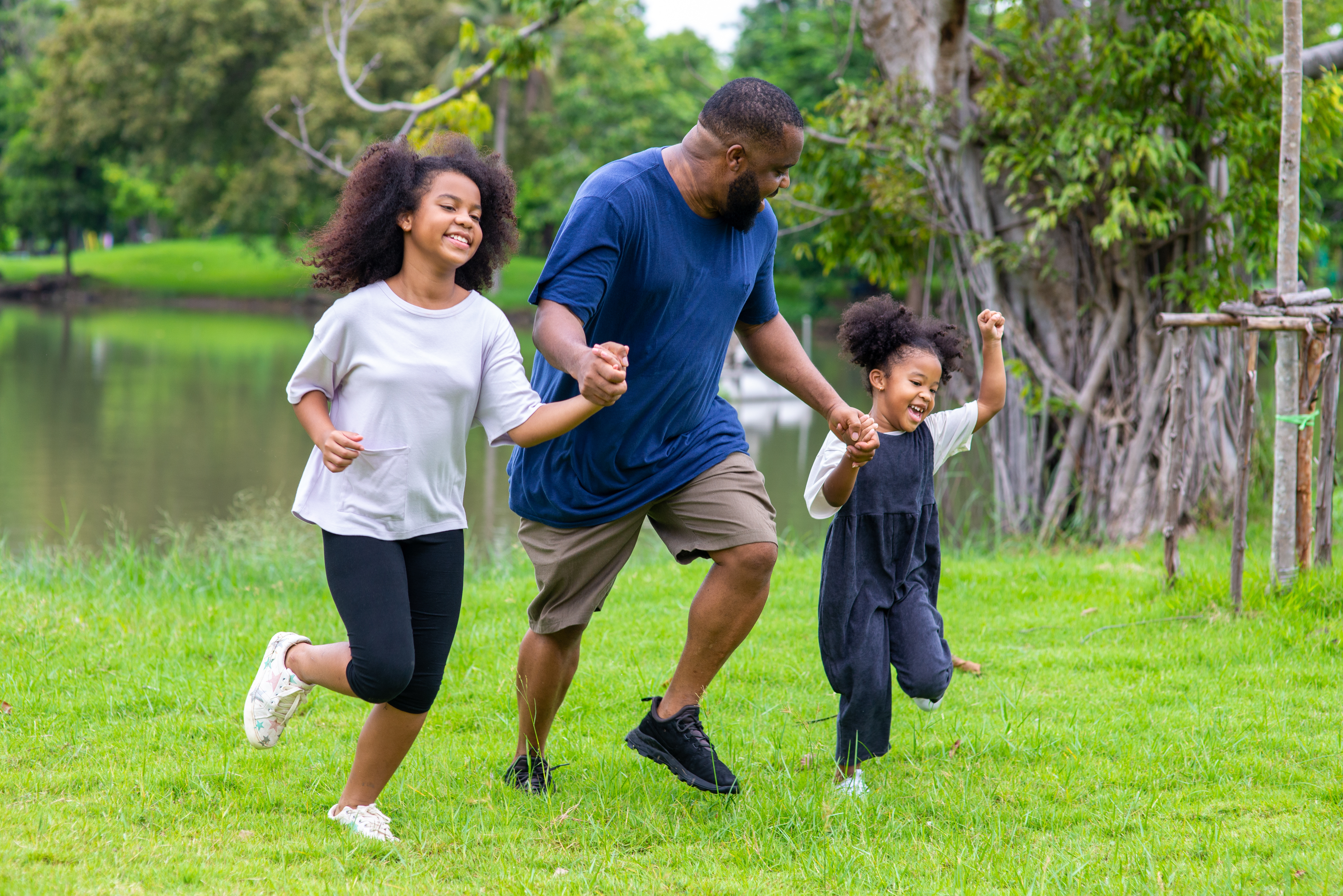 A adult and two children run through grass with trees in the background