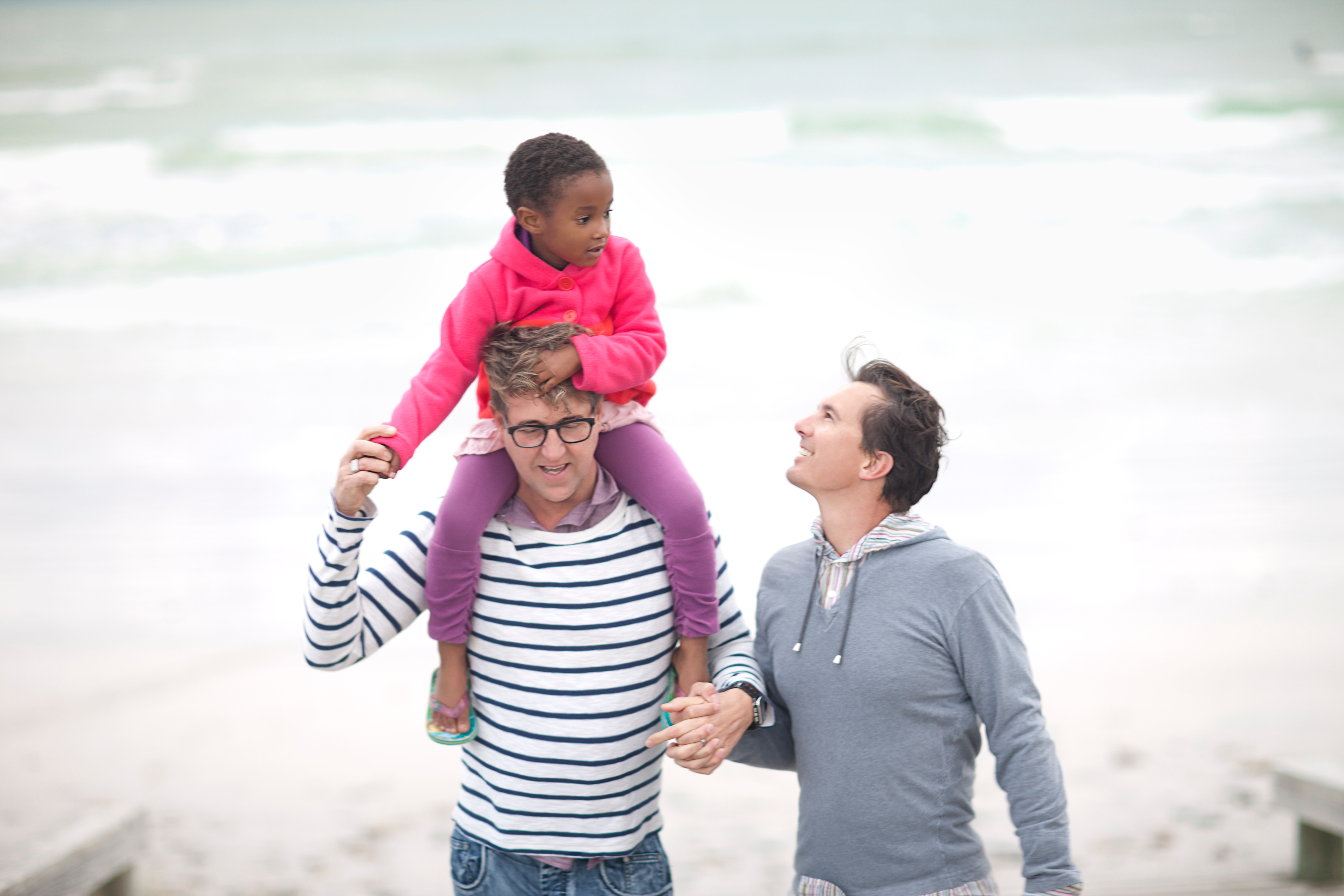 two dads stand on a beach together with a child on their shoulders