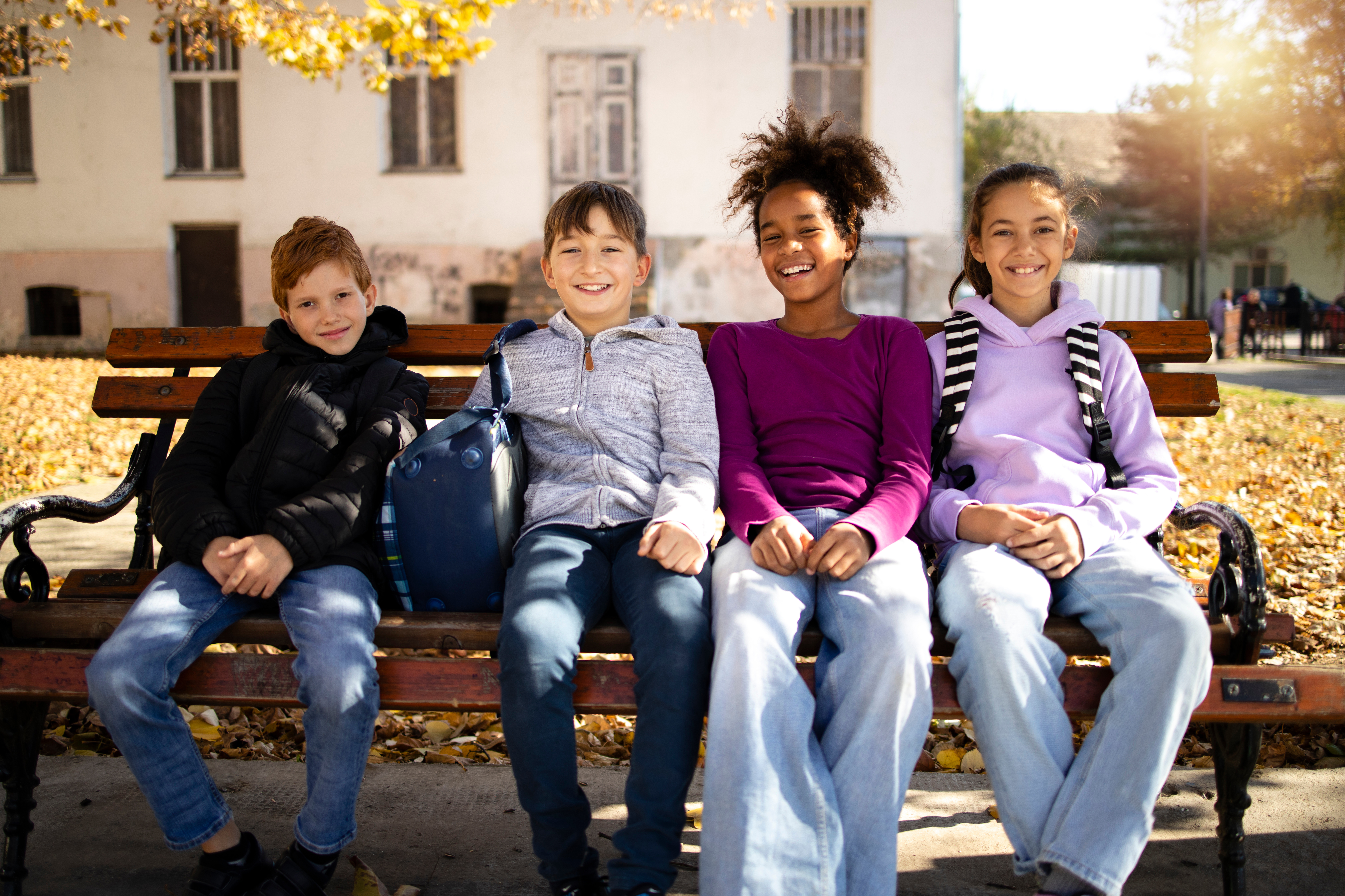 a group of pre-teens sit on a bench together with their backpacks