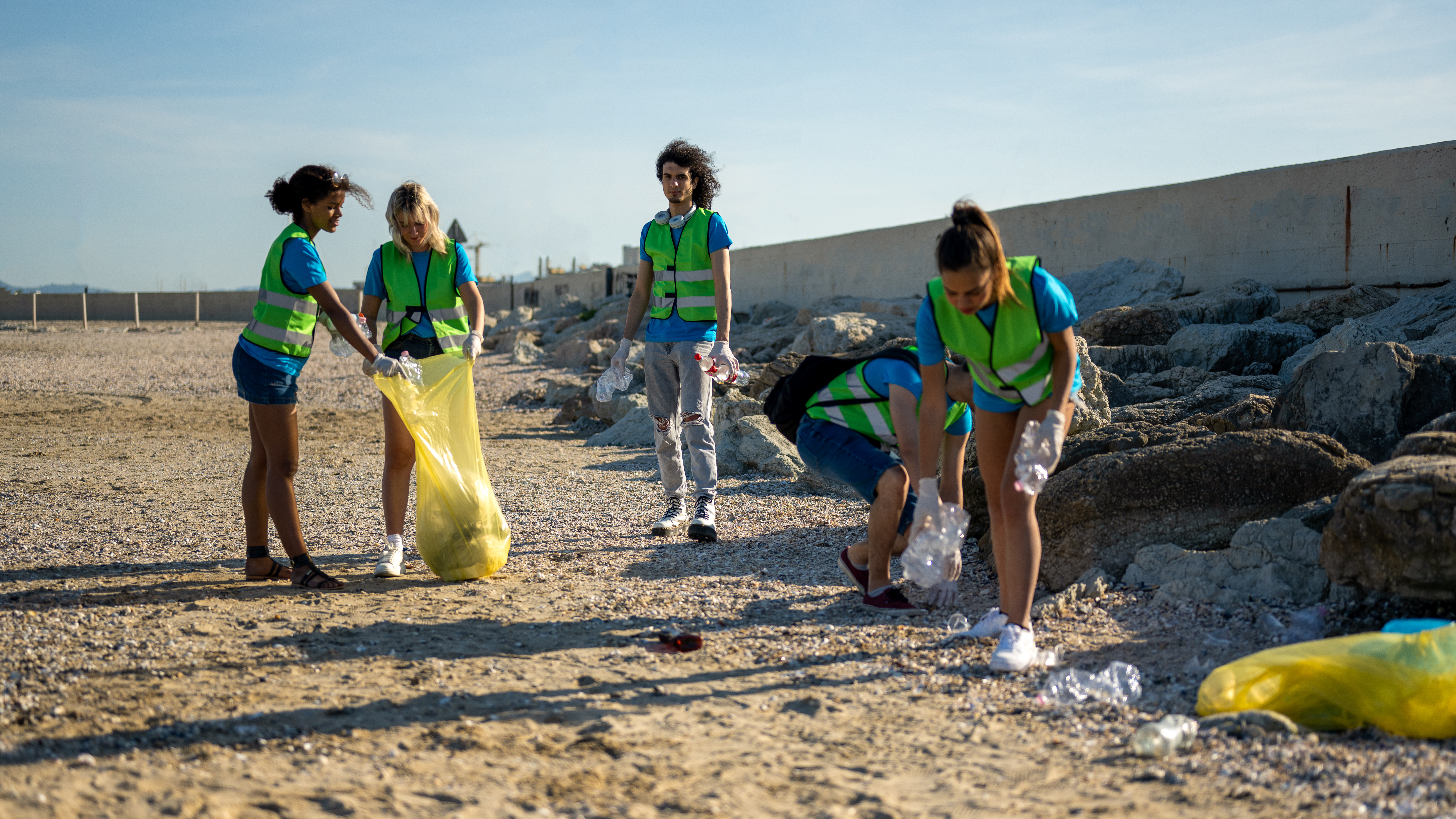 A group of teens in neon vests pick up trash on a beach
