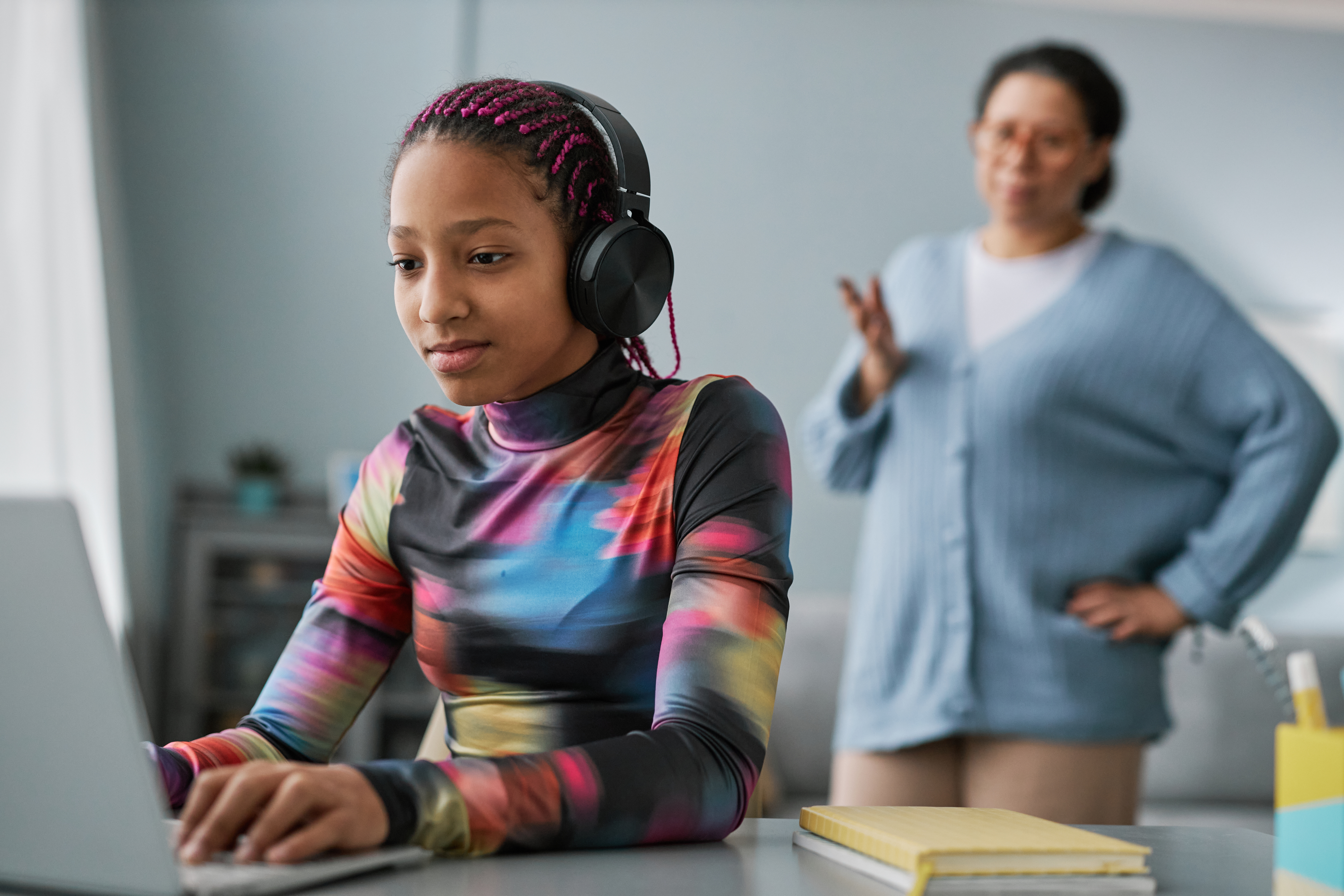 A young person wears headphones and sits at a computer with an adult standing behind