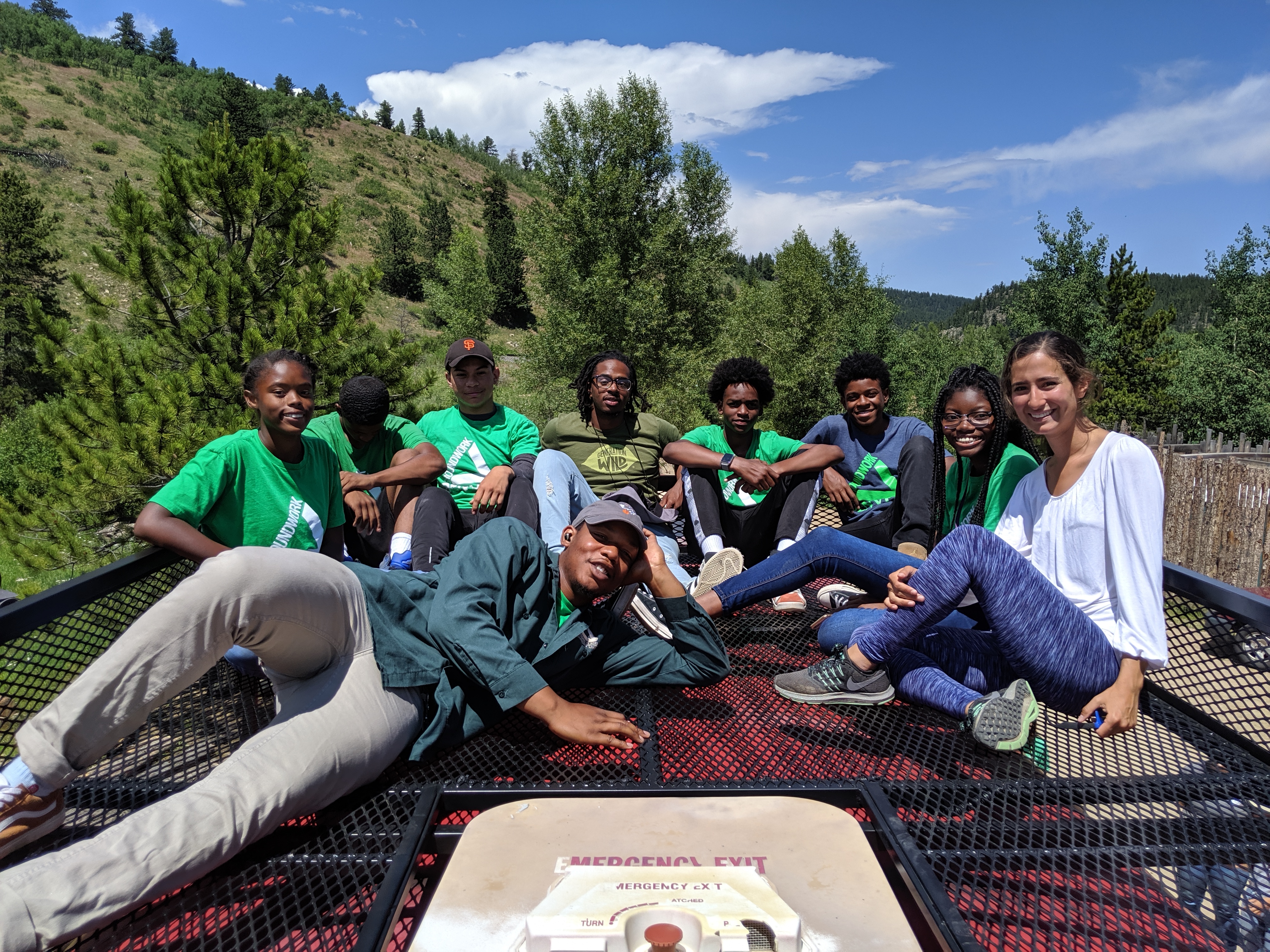 A group of young people and adults sit on top of a bus with mountains in the background