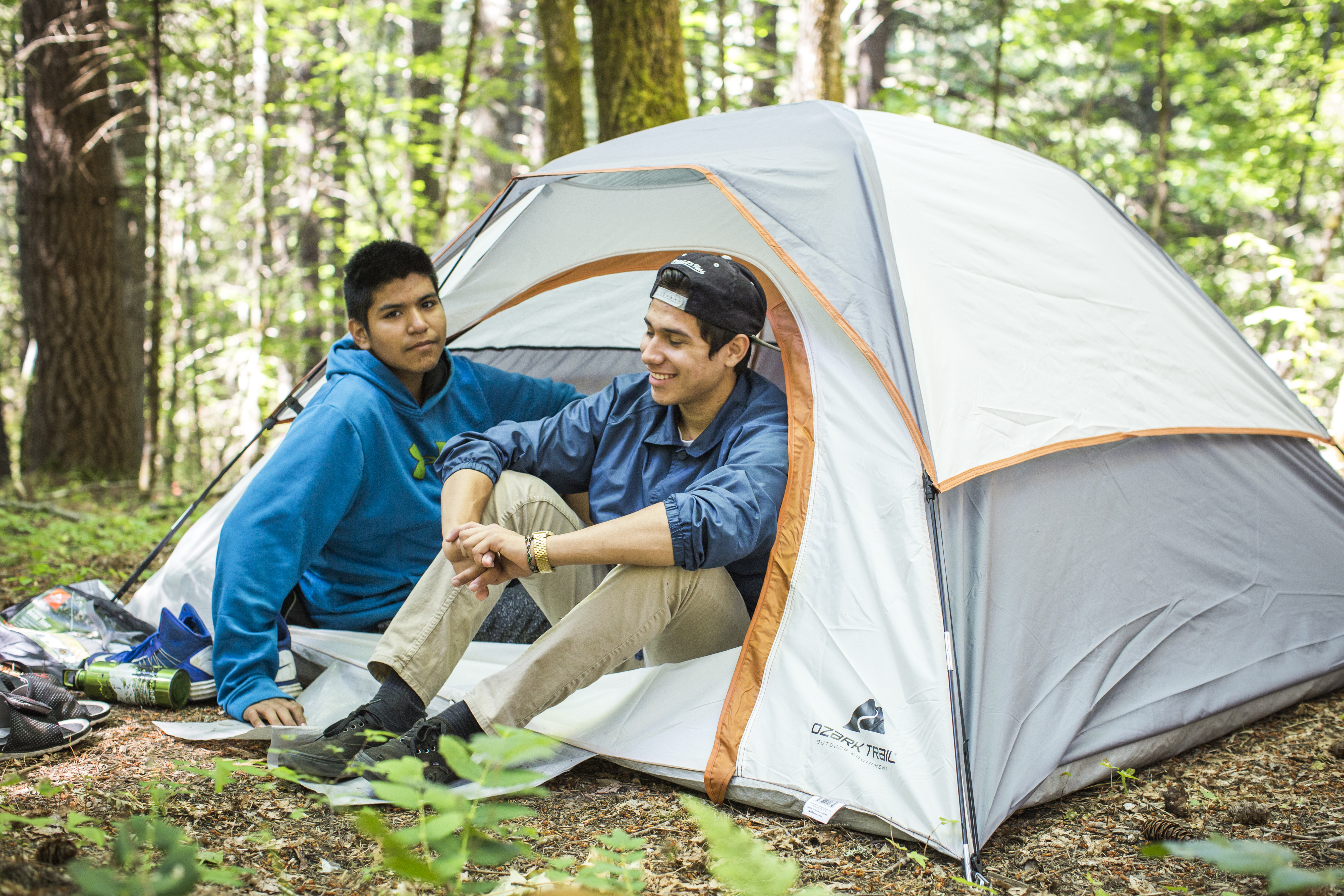 Two adolescent boys sit in a tent, one looking at the camera and one looking into the woods