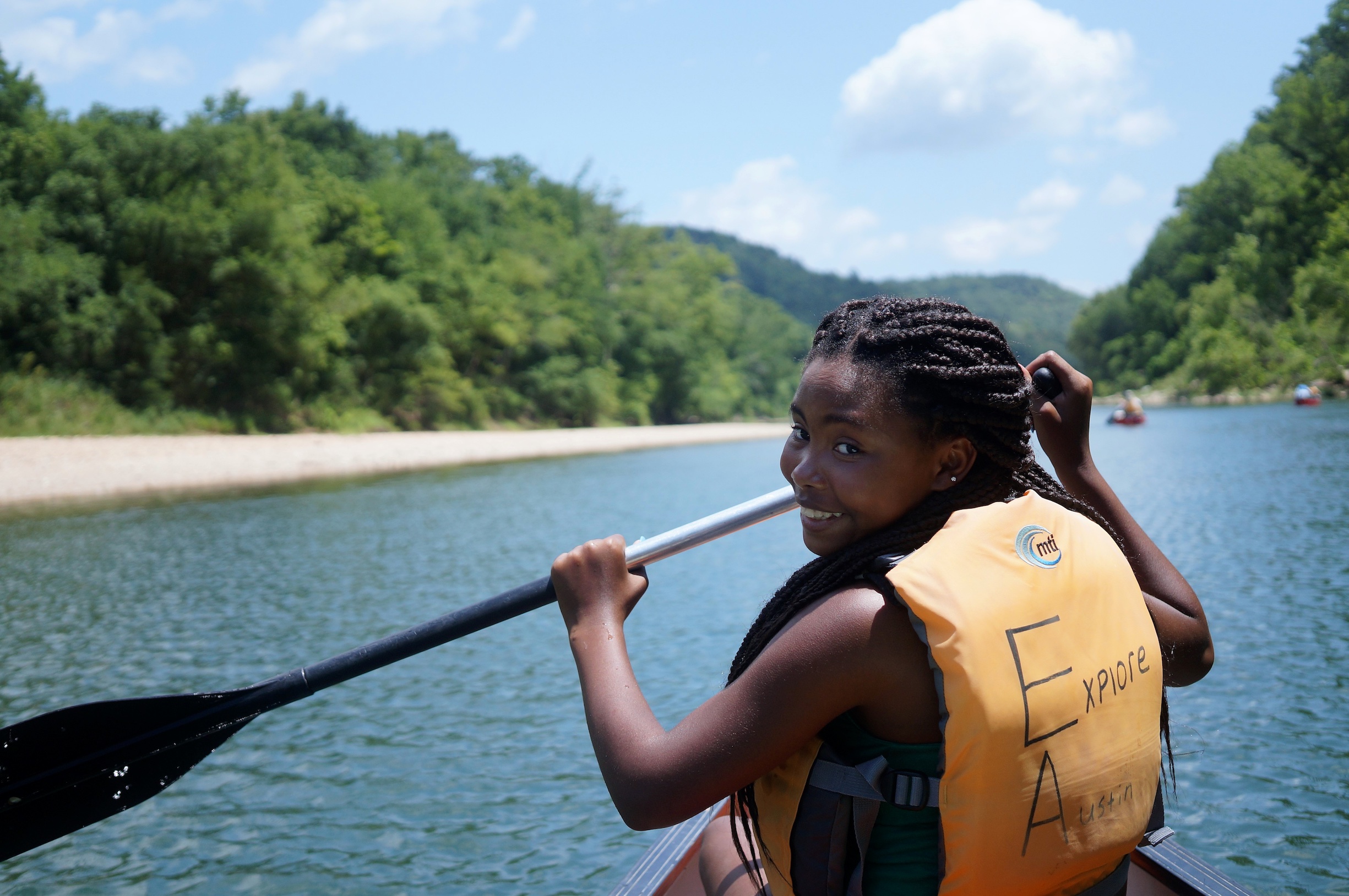 A young woman sits in a boat holding a paddle and looks back at the camera smiling