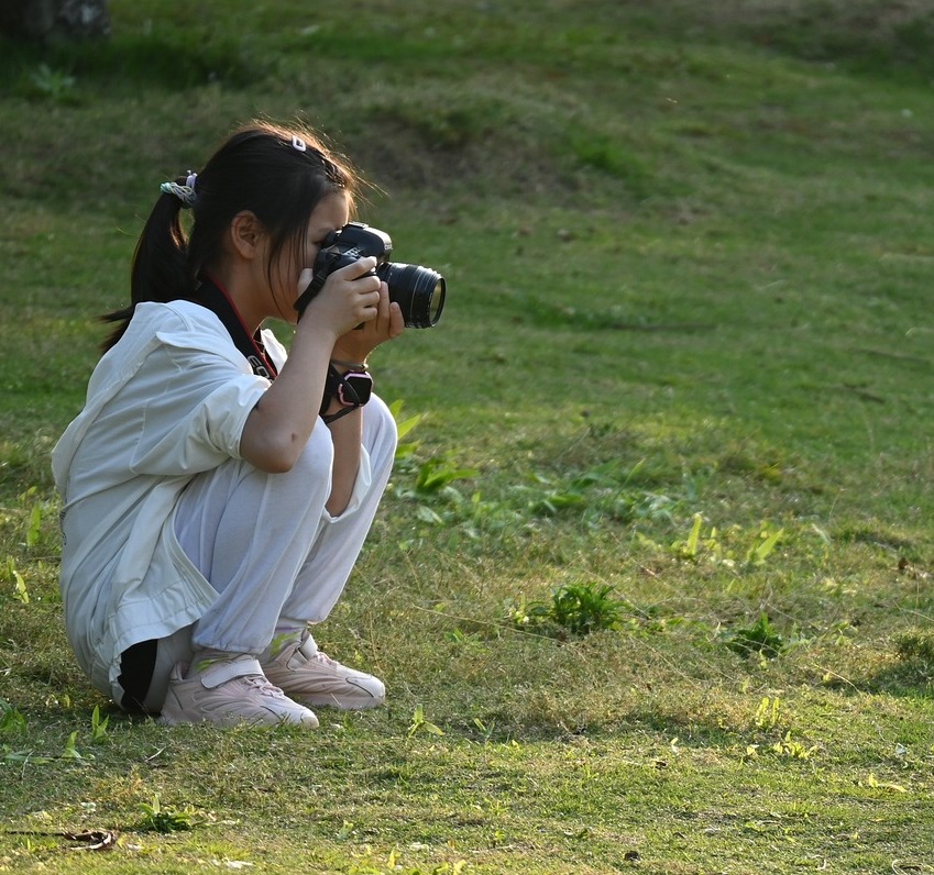 A young person squats on grass taking a picture with a camer